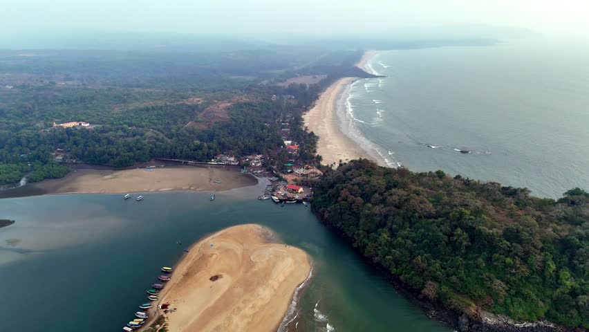 Cinematic aerial view of The LaLiT Golf and Spa Resort Goa, nestled between lush greenery, river backwaters, and the Arabian Sea, with waves rolling along the coastline under a soft hazy sky.