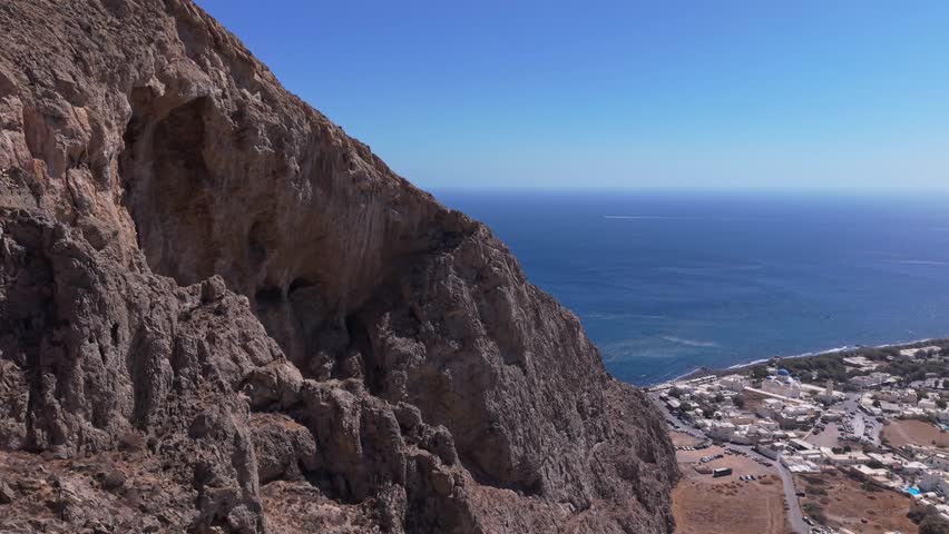 A dramatic drone reveal of the tiny, whitewashed Panagia Katefiani chapel, tucked into a natural rocky alcove high above the black sands of Perissa Beach on the rugged Mesa Vouno cliffs.