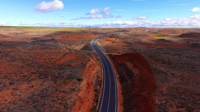 Winding asphalt road cutting through vivid red rocky terrain under a partly cloudy blue sky, stretching toward distant green plains and mountains on the horizon