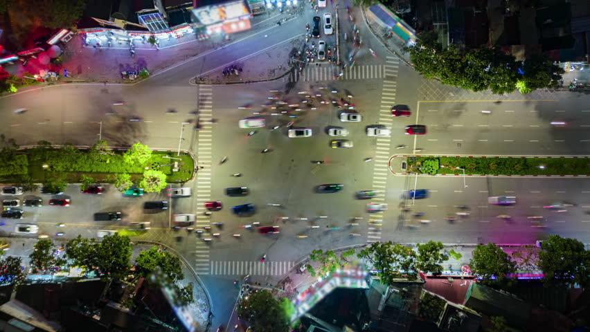 Top down aerial timelapse of busy traffic intersection in Hanoi, Vietnam at night featuring moving cars, motorcycles, crosswalks, city lights, and vibrant urban transportation.