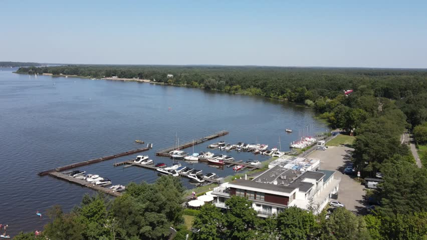Aerial view of a modern marina on a wide lake surrounded by dense green forest with boats docked at piers. Drone shot high angle