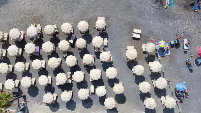 Aerial view of rows of white sun umbrellas and sunbeds on a pebble beach with people relaxing in Imperia, Liguria, Italy.