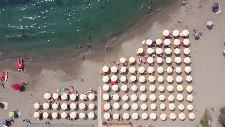 Aerial view of rows of white beach umbrellas on a sandy beach with people swimming in the turquoise sea in Imperia, Liguria, Italy.