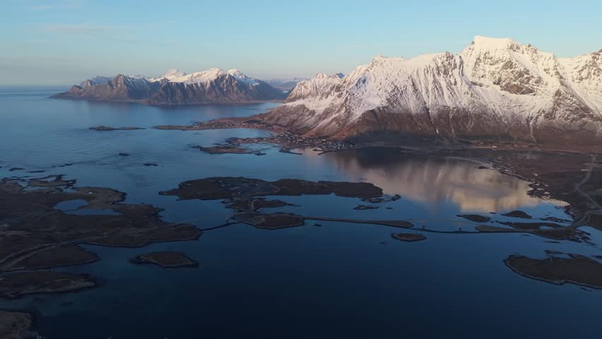 Aerial view shows snow-covered mountains and calm water with islands in northern Norway at dusk.