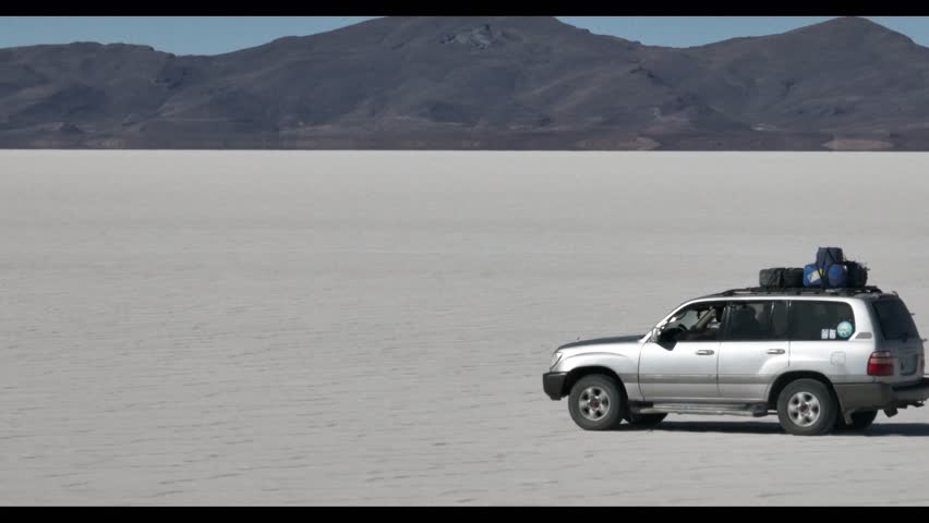 4x4 vehicle driving across the Uyuni salt desert in Bolivia, crossing vast white plains under a clear sky. Minimalist landscape with endless horizon and remote travel adventure.