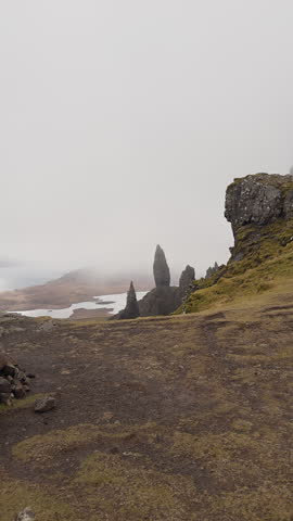 Photographer with backpack facing Old Man of Storr cliffs in misty Scotland highlands landscape. Vertical