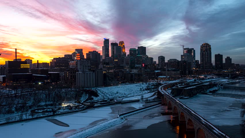 Dynamic aerial motion captures a vibrant sunset over the Minneapolis skyline, highlighting the frozen Mississippi River and the historic Stone Arch Bridge, a captivating winter urban landscape.