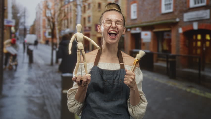 Woman holding wooden mannequin and paintbrush laughing with open mouth on a wet brick street lined with shops; creative joy.