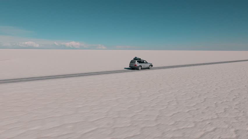 4x4 vehicle driving across the Uyuni salt desert in Bolivia, crossing vast white plains under a clear sky. Minimalist landscape with endless horizon and remote travel adventure.