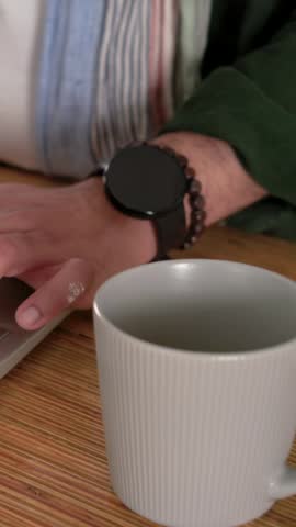 Vertical video: Reaching mug, man in beanie at kitchen bar lifting, sipping, then typing on laptop. Coffee, home, workstation, keyboard, screen, minimal, vertical video