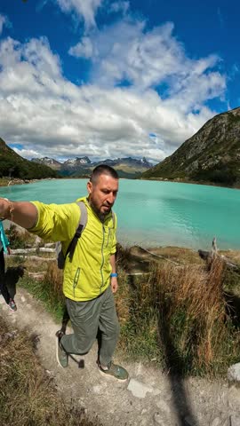 A point of view shot shows a man and woman hiking on a trail. They are walking along the turquoise waters of Laguna Esmeralda, Ushuaia, Argentina.