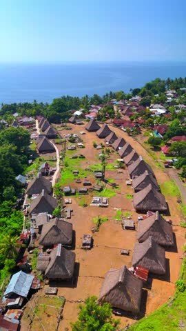 Aerial view of Kampung Adat Nggela traditional village with thatched roofs and lush greenery overlooking the ocean in Wolojita, East Nusa Tenggara, Indonesia.