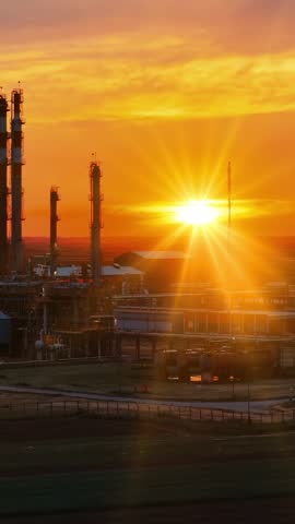 Cinematic aerial view of a petrochemical plant at sunset. Industrial towers, pipelines, and refinery structures stand against a glowing orange sky as warm sunlight reflects across the facility
