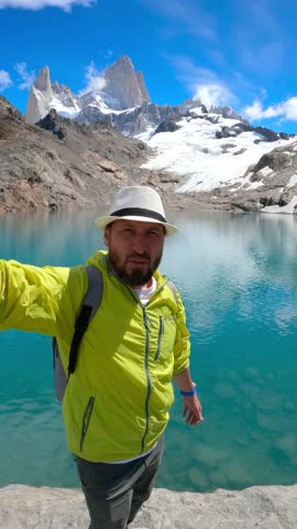 Middle-aged man wearing lime green jacket and hat taking selfie at Laguna de los Tres with Mount Fitz Roy in the background, Patagonia, Argentina.