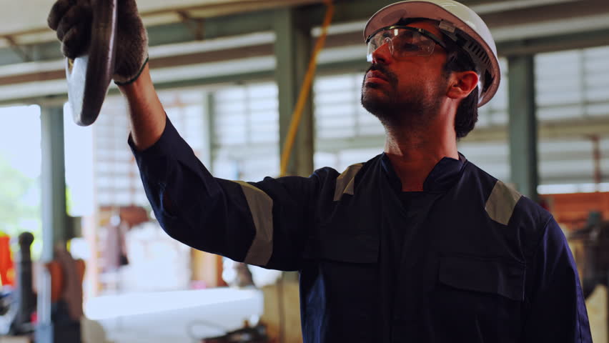 Serious focused man factory worker wearing safety glasses and white helmet inspecting heavy industrial manufacturing equipment with large metal crane hook during indoor operation