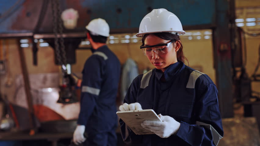 Focused female engineer in white hardhat and protective glasses using digital tablet for inspecting heavy machinery equipment with male coworker inside industrial manufacturing factory
