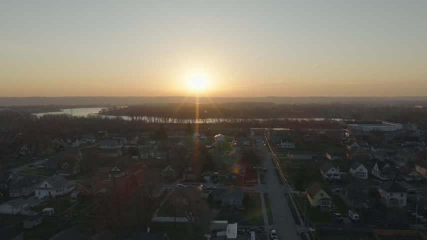 Aerial sideways flight over Omaha Nebraska suburbs at sunset captures glowing sun touching horizon above winding Missouri River and tree-lined residential neighborhoods