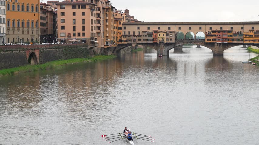 Competitive rowers glide across the Arno River in Florence, Italy, with Ponte Vecchio rising behind. Overcast daylight creates soft reflections and highlights iconic Renaissance architecture.