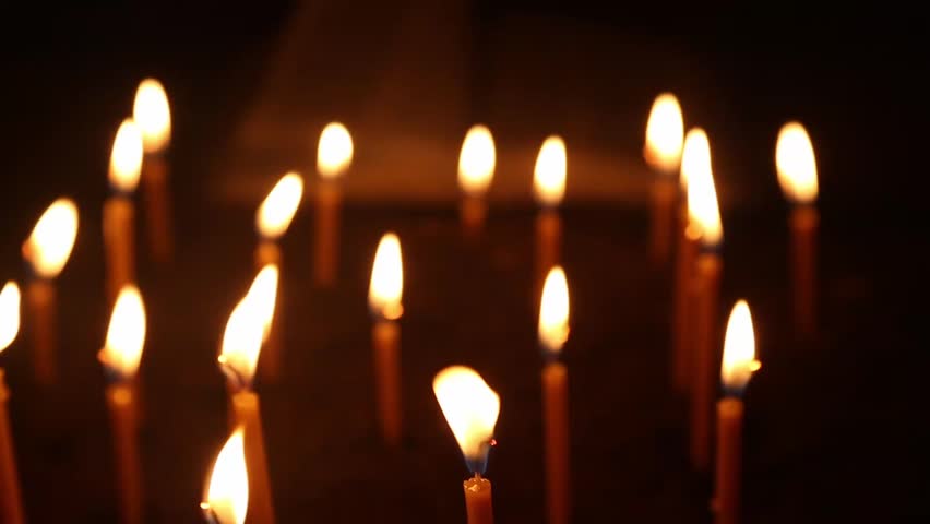 Rows of burning church candles in a dim religious interior, the flames flickering and swaying in the gentle movement of air, with a blurred background showing a book and the turning of pages