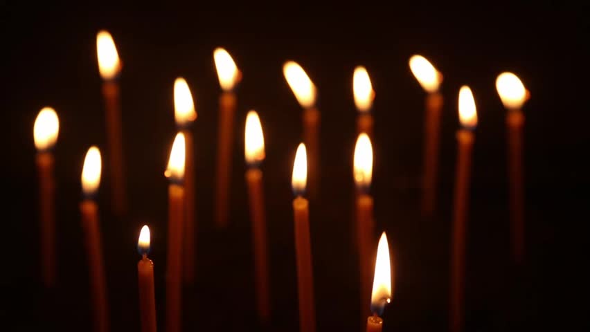 A human hand uses a candle to light one tall church candle among many glowing candles against a dark background.
