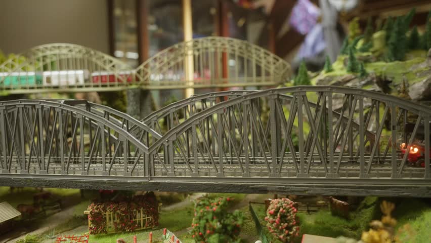 A train in a miniature landscape enters the frame from the right and travels across an iron bridge toward the left in a tracking shot