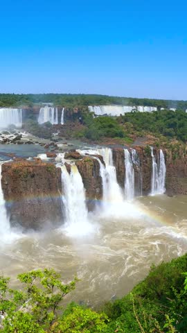 Cascading waterfalls flow over rocky cliffs beside a rainbow and lush green forest at Iguazu Falls on the border of Argentina and Brazil.