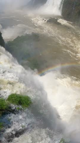 High angle view of water cascading over rocky cliffs with rising mist and a rainbow at Iguazu Falls, Argentina and Brazil.