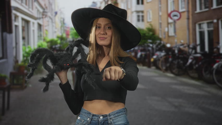 Young woman holding a large black spider plush and showing bare midriff on a cobbled street lined with bicycles; playful spooky fun.