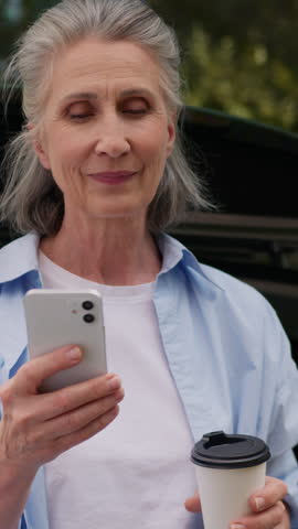 Cheerful Caucasian retired old senior woman calling talking mobile phone with family drinking coffee sitting in car auto trunk at nature travel aged mature senior female talk laugh near automobile