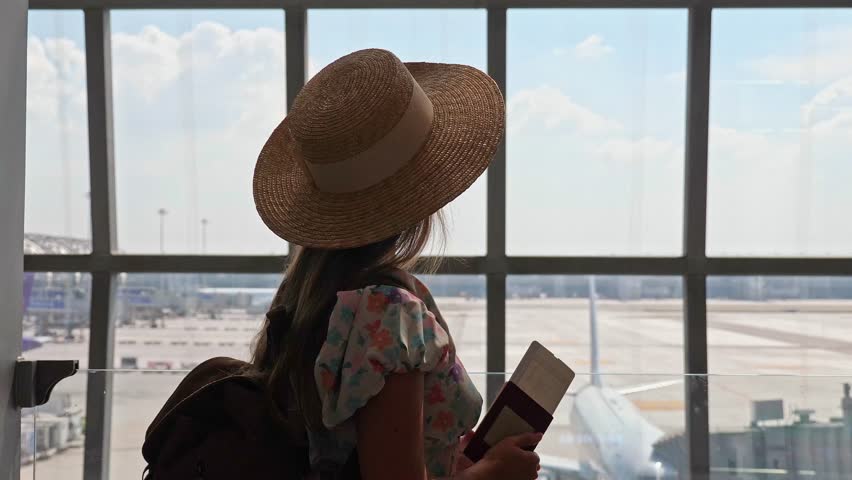 Woman in straw hat holding passport and boarding pass looking at aircraft through glass window, airport terminal departure lounge, international travel, summer vacation background, slow motion