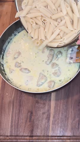 Vertical cooking footage showing cooked penne pasta being added into a large frying pan containing creamy milk sauce and chicken breast pieces. Fresh sliced mushrooms are added next before the ingredients are stirred with a wooden spatula. The pan is then covered with a glass lid and in the final scene the lid is lifted as steam escapes revealing creamy chicken mushroom penne Alfredo pasta gently bubbling inside the skillet. Suitable for homemade pasta recipes, creamy comfort food videos, and cooking tutorial content.
