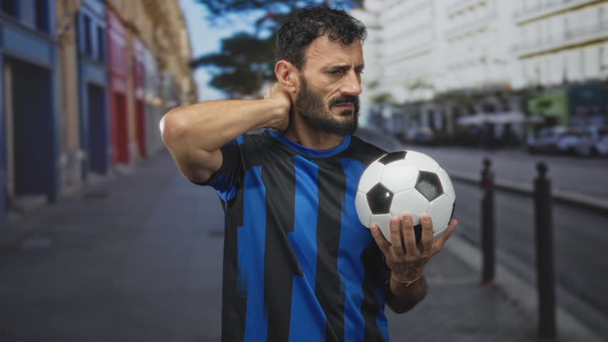 Man holding a soccer ball and rubbing his neck on a city street lined with buildings and bollards, wearing a blue striped uniform; discomfort resilience.