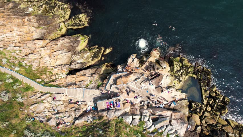 Aerial view of Vico Bathing Place with people gathered on the rocky shoreline and swimming in the sea Killiney, County Dublin, Ireland.