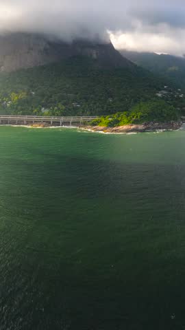 Aerial view of a coastal highway bridge stretching along rocky cliffs and lush green mountains under heavy clouds in Rio de Janeiro, Brazil.