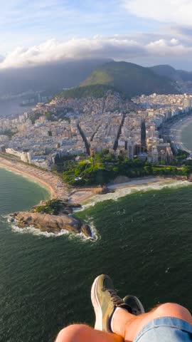 POV view of a young man paragliding over the urban coastline, Arpoador rock, and the Atlantic Ocean in Rio de Janeiro, Brazil.