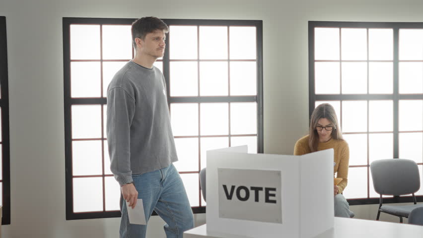 Man places ballot into a clear vote box in a polling building while a woman sits checking a clipboard nearby, us flags and a vote booth visible; civic duty.