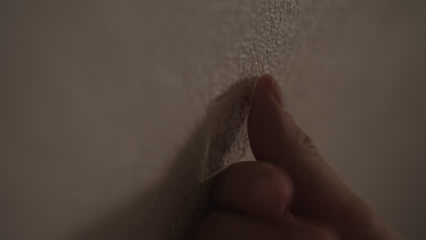 Close-up of fingers peel thin strip of flaking paint or wallpaper from textured wall. Detail shot conveys anxiety, boredom, deconstruction in dark interior of room. Shooting in slow motion.