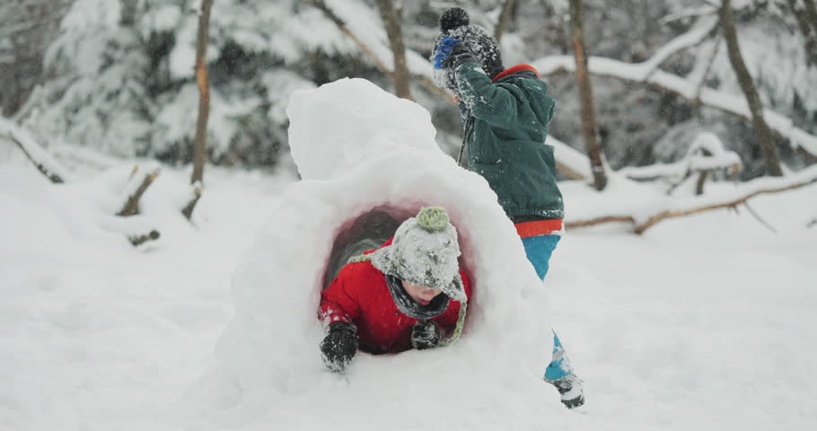 Two children bundled in winter gear walking and playing together in deep snow next to a giant snowball