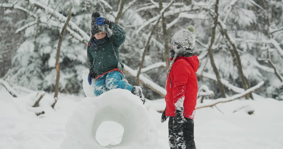 Two children standing next to a large, hollowed-out snow tunnel in a winter forest setting
