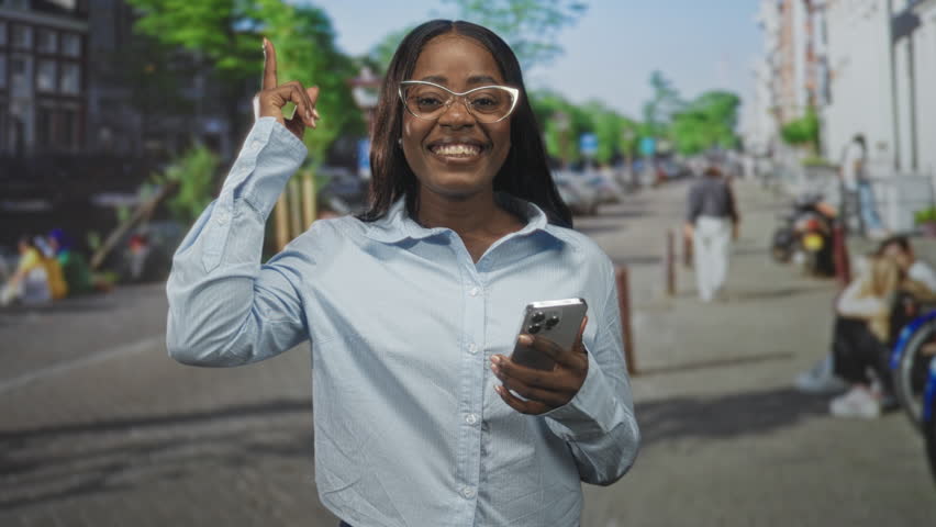Woman holding smartphone and points index finger up on a busy city street while smiling and wearing glasses; joy connection.