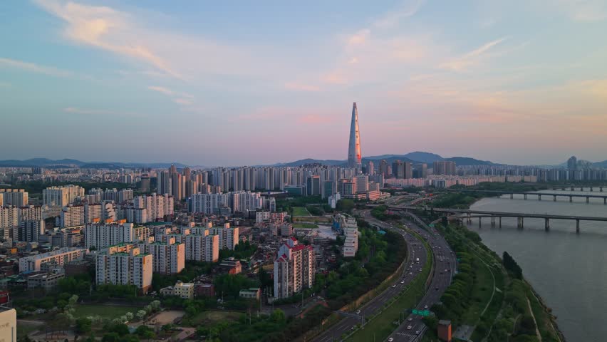 Aerial forward push toward glowing Lotte World Tower at spring sunset over Olympic-daero expressway traffic and Songpa district apartment cityscape