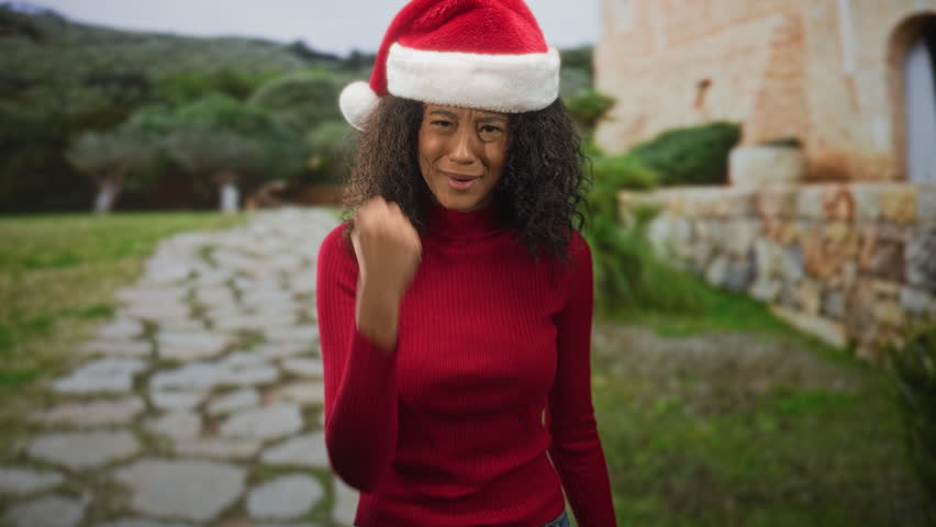 Teenage woman with curly hair and santa hat raises clenched fist, mouth open and eyes wide, standing on a stone path by a building; defiance.