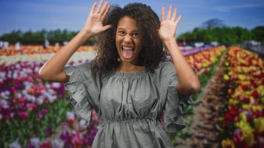 Teenage african american woman with curly hair crosses arms and pouts wearing gingham dress in studio with tulip backdrop; playful mischief.