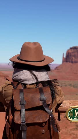Young woman with a backpack and hat enjoying the iconic western landscape of monument valley, turning to the camera and smiling on a sunny day during her amazing american road trip vacation