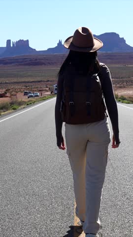 Courageous female hiker with a backpack and hat walking down a long, empty desert highway toward the iconic sandstone buttes in monument valley, enjoying the scenic beauty of the american west