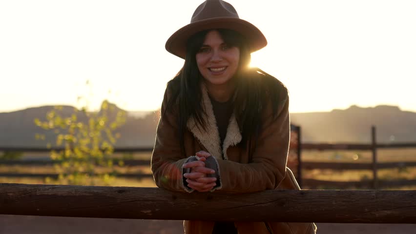 Happy young woman wearing a cowboy hat and leaning on a rustic wooden fence while enjoying the warm, golden sunlight of sunrise in the iconic american west landscape of monument valley