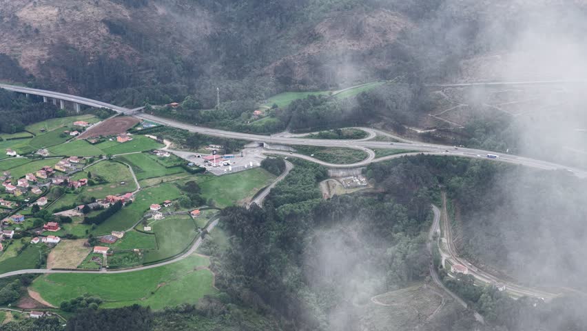 Aerial view of a highway bridge and road interchange surrounded by green fields and rolling fog in Asturias, Asturias, Spain.