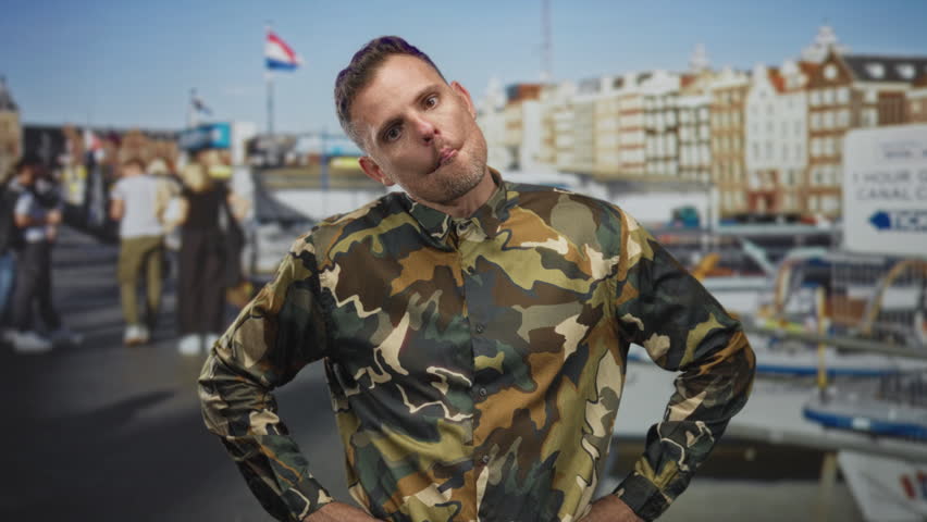 Man soldier in camouflage uniform with hands on hips and pursed lips, standing before canal buildings and boats on a street; confidence duty.
