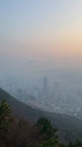 Panoramic panning of Busan cityscape at sunset from a mountain hilltop in South Korea. Dense urban skyline with high-rise residential and commercial skyscrapers surrounded by mountains under a hazy pink and orange sky