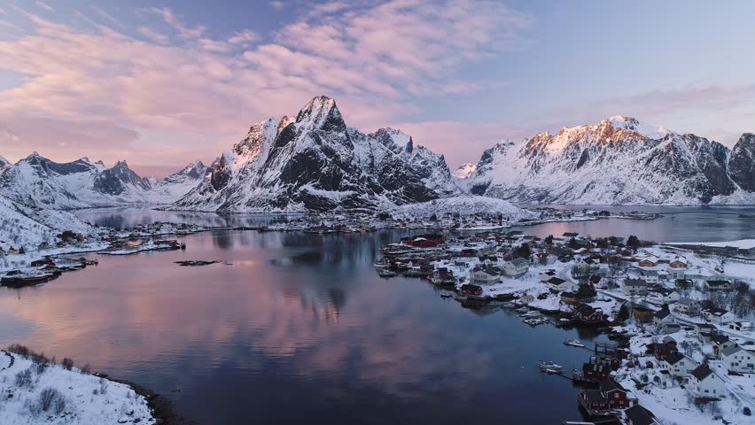 Aerial view of the snow-covered Lofoten mountains reflecting in the calm water of the fjord at sunset in Reine, Nordland, Norway.